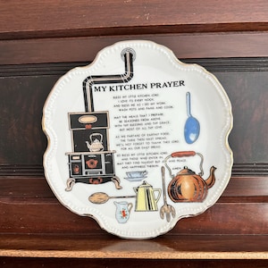 Puede incluir: Un plato decorativo de cerámica blanca con borde dorado festoneado que presenta una ilustración negra de una estufa de leña antigua, una tetera de cobre, una cuchara azul, una cafetera amarilla y una jarra pequeña. El texto dice MY KITCHEN PRAYER.