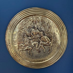 May include: A round, gold-colored metal relief depicting a scene with three figures seated around a table. The relief has a detailed, textured design with a ridged border. The background is a dark blue.