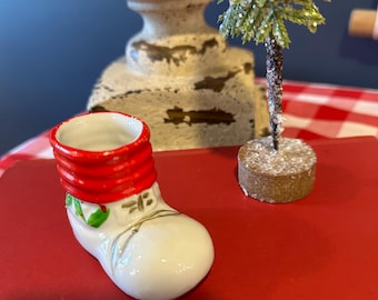Puede incluir: Una pequeña bota navideña de cerámica con la parte superior roja acanalada y el cuerpo blanco. La bota está decorada con hojas de acebo verdes y detalles dorados. También hay un árbol de Navidad en miniatura con agujas verdes brillantes.