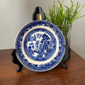 Puede incluir: Un plato de cerámica azul y blanco con un diseño Willow, exhibido en un soporte de madera negro. El plato presenta una escena de paisaje detallada con árboles, edificios y un puente. El plato tiene aproximadamente 20 cm de diámetro.