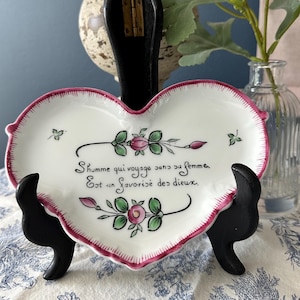 May include: Heart-shaped white ceramic dish with a pink border and floral design. The dish features a French quote in black script. The dish is supported by a black stand, and is a decorative item.