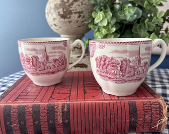 May include: Two white ceramic teacups featuring a detailed red transferware print of a cathedral and landscape scene. Each cup has a curved handle and a scalloped rim design. The cups are positioned on top of a red and black vintage hardcover book.