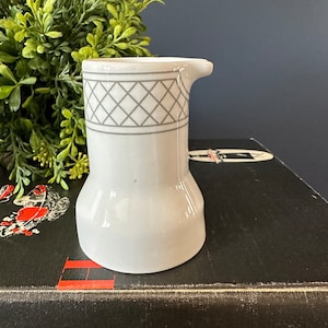 以下が含まれることがあります： 上部にグレーのダイヤモンド模様が施された白いセラミック製のクリーマー。クリーマーは小さな注ぎ口と丸みを帯びた底を持っています。