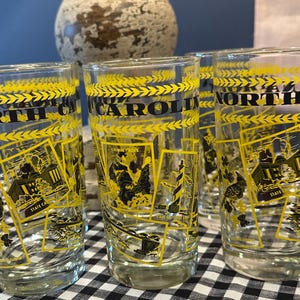 May include: Set of clear drinking glasses with yellow and black designs. Each glass features the words "NORTH CAROLINA" and illustrations of state landmarks. The glasses are on a black and white checkered surface.