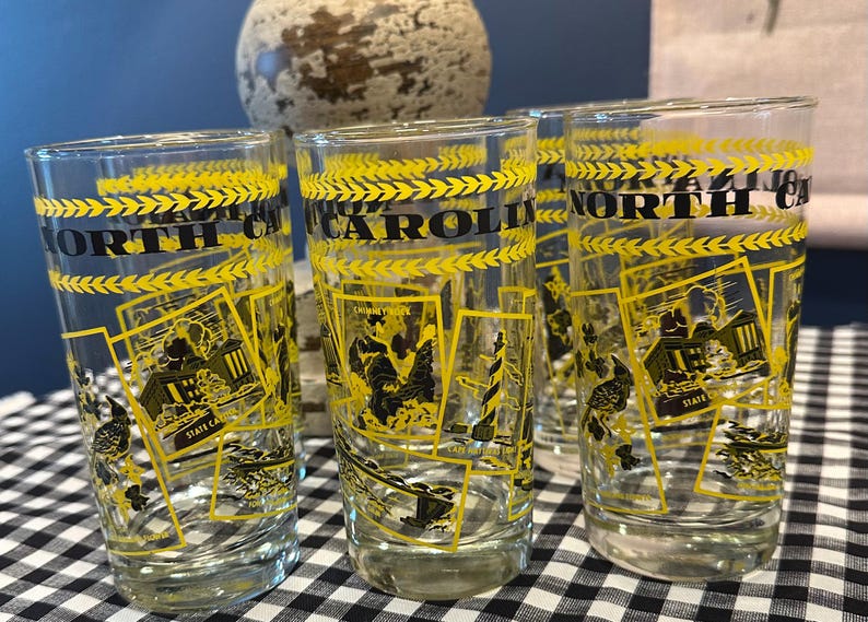 May include: Set of clear drinking glasses with yellow and black designs. Each glass features the words "NORTH CAROLINA" and illustrations of state landmarks. The glasses are on a black and white checkered surface.