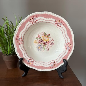 May include: A white ceramic bowl with a pink scalloped rim and floral design. The bowl features a central bouquet of yellow, purple, and burgundy flowers. The rim has a repeating pattern of small flowers and leaves. The bowl is displayed on a black stand.