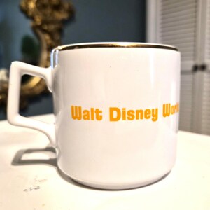 Puede incluir: Taza de cer&aacute;mica blanca con borde dorado y asa cuadrada. La taza presenta el texto "Walt Disney World" en naranja. Un recuerdo cl&aacute;sico para caf&eacute; o t&eacute;.