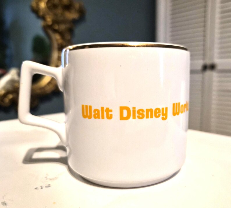 Puede incluir: Taza de cer&aacute;mica blanca con borde dorado y asa cuadrada. La taza presenta el texto "Walt Disney World" en naranja. Un recuerdo cl&aacute;sico para caf&eacute; o t&eacute;.