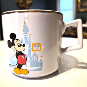 Puede incluir: Taza de cer&aacute;mica blanca con borde dorado que presenta una imagen de dibujos animados de Mickey Mouse con su atuendo cl&aacute;sico, frente a un castillo azul. La taza tiene un asa cuadrada y un logotipo de Disney.