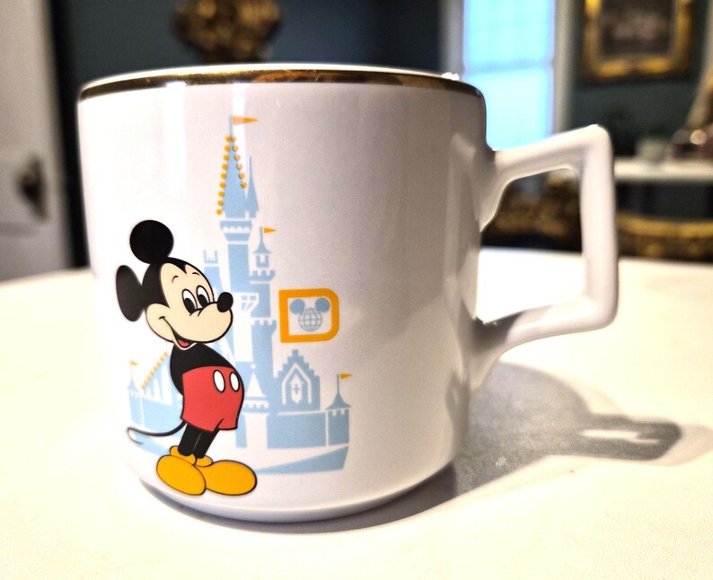 Puede incluir: Taza de cer&aacute;mica blanca con borde dorado que presenta una imagen de dibujos animados de Mickey Mouse con su atuendo cl&aacute;sico, frente a un castillo azul. La taza tiene un asa cuadrada y un logotipo de Disney.