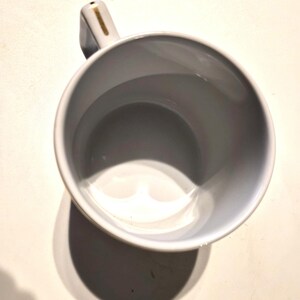 Puede incluir: Una taza de cer&aacute;mica blanca con interior gris y asa. El asa tiene un acento dorado. La taza est&aacute; vac&iacute;a y proyecta una sombra sobre una superficie blanca.