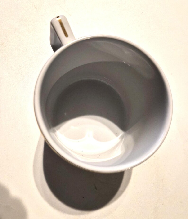 Puede incluir: Una taza de cer&aacute;mica blanca con interior gris y asa. El asa tiene un acento dorado. La taza est&aacute; vac&iacute;a y proyecta una sombra sobre una superficie blanca.