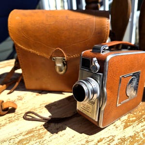 May include: An antique brown leather camera and matching case sit on a wooden surface. The camera has a silver lens and trim, with a label that reads "Real Air Magazine 8". The case has a flap closure and a shoulder strap.