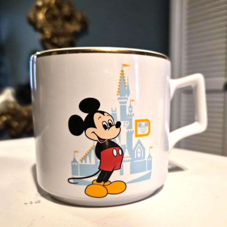 Puede incluir: Taza de cer&aacute;mica blanca con borde dorado, con una imagen de dibujos animados de Mickey Mouse y un dise&ntilde;o de castillo azul claro. La taza tiene un asa cuadrada.