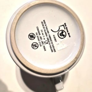 Puede incluir: Taza de cer&aacute;mica blanca con asa, con el logotipo del 50 aniversario de Walt Disney World y texto. La parte inferior de la taza incluye detalles de fabricaci&oacute;n, informaci&oacute;n sobre el material y la capacidad. La taza es de gres y contiene 312 ml.