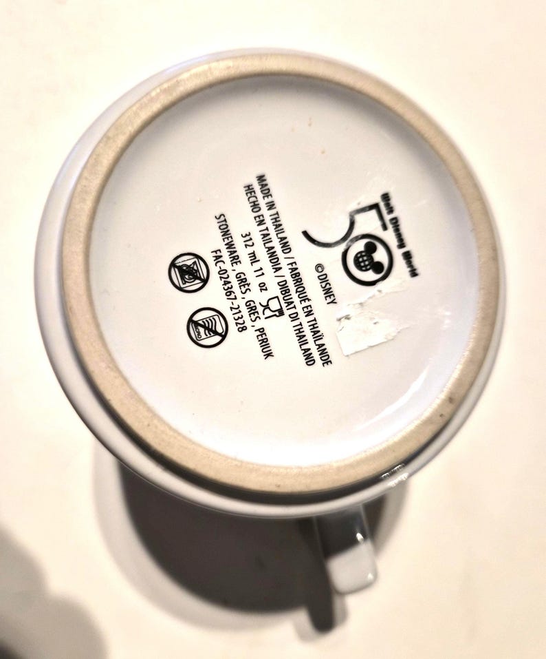 Puede incluir: Taza de cer&aacute;mica blanca con asa, con el logotipo del 50 aniversario de Walt Disney World y texto. La parte inferior de la taza incluye detalles de fabricaci&oacute;n, informaci&oacute;n sobre el material y la capacidad. La taza es de gres y contiene 312 ml.