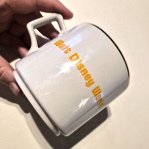 Puede incluir: Taza de cer&aacute;mica blanca con el texto "Walt Disney World" en letras naranjas. La taza tiene un borde negro y un asa cuadrada. La taza est&aacute; sujeta por una mano.