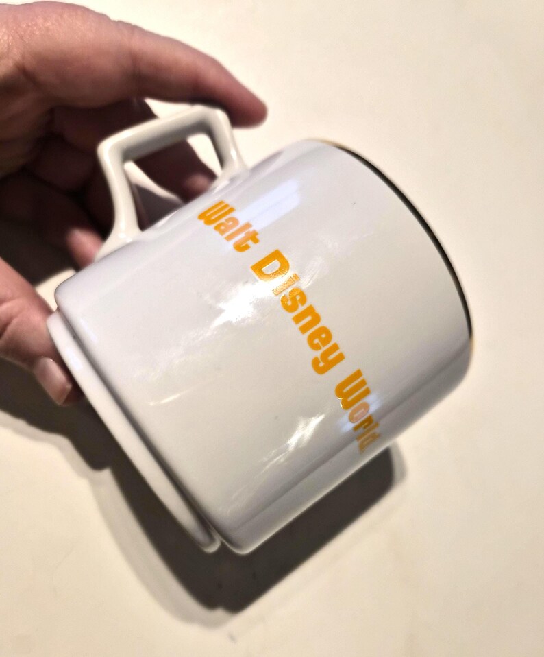 Puede incluir: Taza de cer&aacute;mica blanca con el texto "Walt Disney World" en letras naranjas. La taza tiene un borde negro y un asa cuadrada. La taza est&aacute; sujeta por una mano.