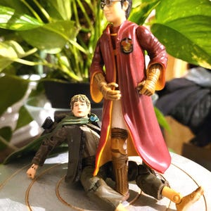 May include: Two action figures, one in a red and gold robe with glasses, and the other in a green and brown outfit, are posed on a light-colored surface. The figure in the robe is standing over the other figure.