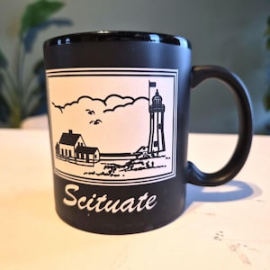 May include: Black ceramic mug featuring a white rectangular design with a lighthouse, house, and the word "Scituate". The mug has a curved handle and is suitable for hot or cold beverages.
