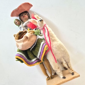 May include: A handcrafted figurine depicting a woman in traditional attire, carrying a child and a clay pot, accompanied by a llama. The woman wears a pink hat and a green, white, and yellow striped garment. The llama has a white body and a pink and white lead.