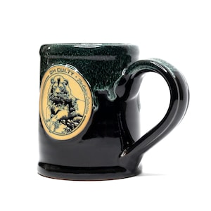 May include: A black ceramic mug with a large handle. The mug has a dark green rim and a circular emblem with a lion illustration and the text "OH CULTY" and "The Cabija Collection".