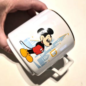 Puede incluir: Taza de cer&aacute;mica blanca con Mickey Mouse con su atuendo cl&aacute;sico, con un dise&ntilde;o de castillo azul claro. La taza tiene un borde dorado y un asa blanca. Un cl&aacute;sico de colecci&oacute;n de Disney.