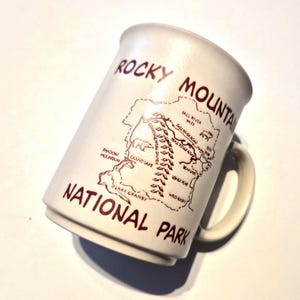 May include: Cream-colored ceramic mug featuring a map of Rocky Mountain National Park. The mug has a handle and is printed with the words "Rocky Mountain National Park" in a dark red color. The map includes roads and landmarks.