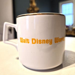 Puede incluir: Taza de cer&aacute;mica blanca con borde dorado y asa cuadrada. Las palabras "Walt Disney World" est&aacute;n impresas en naranja en el lateral. La taza est&aacute; sobre una superficie blanca.