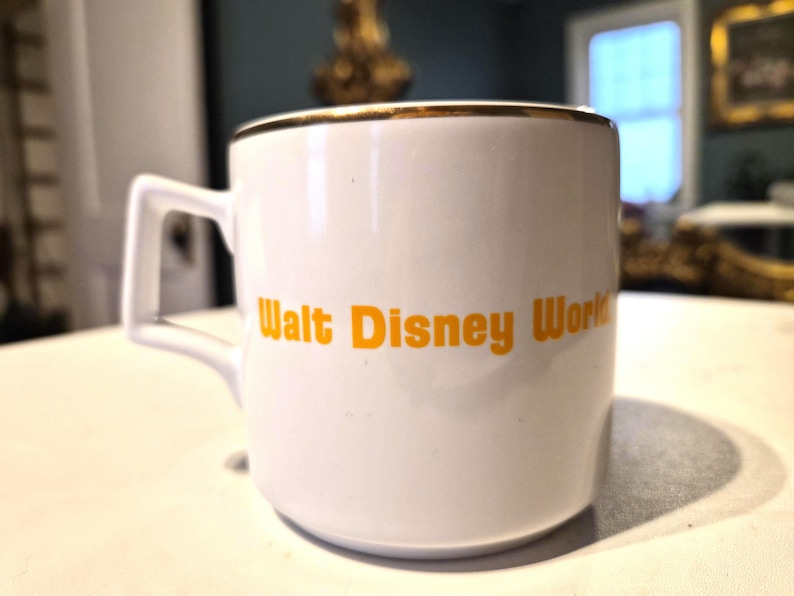 Puede incluir: Taza de cer&aacute;mica blanca con borde dorado y asa cuadrada. Las palabras "Walt Disney World" est&aacute;n impresas en naranja en el lateral. La taza est&aacute; sobre una superficie blanca.