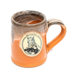 May include: Orange ceramic mug with a brown rim and handle. A circular emblem on the front features a tiger illustration and the text "OH CULTY" and "The Cobija Collection". The mug has a glossy finish and a curved handle.