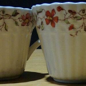 May include: Two white ceramic teacups with a floral pattern in shades of pink and brown. The cups have a fluted design and a handle.