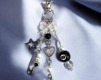 May include: A silver charm keychain with a variety of charms, including a bow, a star, a heart, a safety pin, and a black and white 8-ball. The charms are attached to a silver chain.