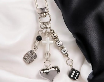May include: A silver keychain featuring an assortment of charms, including a cross, a heart, a dice, and letter beads spelling "OKJ". The keychain also includes a small silver tag with the inscription "Handmade with love, Bless Button Boutique, est. 2017".