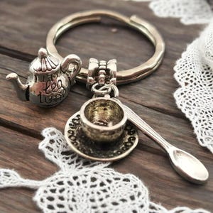 Puede incluir: Llavero con un charm de plata que representa una tetera con la palabra "té" en ella, una taza de té pequeña y un platillo, y una cuchara. El charm está unido a un llavero de plata.