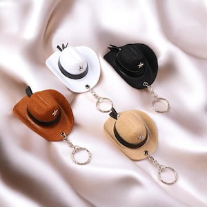 May include: Four cowboy hat keychains in different colors: brown, white, black, and beige. Each hat has a silver star on the front and a silver chain attached to a key ring.