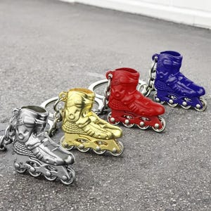Könnte beinhalten: Vier Rollschuh-Schlüsselanhänger in verschiedenen Farben: Silber, Gold, Rot und Blau. Jeder Schlüsselanhänger zeigt ein detailliertes Rollschuh-Design mit Rädern und einem Metallring.