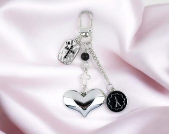 May include: Silver keyring with a heart, cross, and a black and silver charm with a design. The keyring is on a white satin background.
