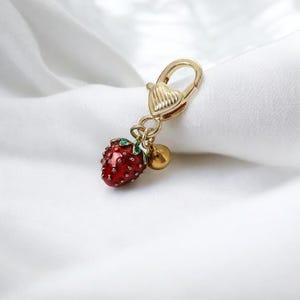 May include: Gold-tone keychain with a heart-shaped charm and a red enamel strawberry charm. The strawberry charm has green leaves and a gold-tone bell.