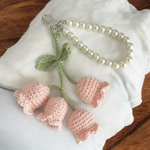 May include: A pink crocheted flower keychain with green leaves and a pearl chain. The keychain is attached to a silver metal clasp.
