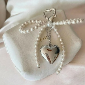 May include: A silver heart-shaped charm with a pearl accent hangs from a silver chain. The chain is attached to a silver heart-shaped clasp. The charm is surrounded by a white pearl necklace.