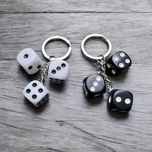 Puede incluir: Dos llaveros con dijes de dados. Un llavero tiene tres dados blancos con puntos negros, y el otro llavero tiene tres dados negros con puntos blancos.