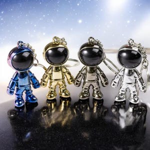Puede incluir: Cuatro llaveros de astronauta en diferentes colores: azul, dorado, plateado y negro. Los llaveros de astronauta llevan trajes espaciales y tienen viseras negras.