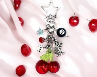 May include: A silver charm keychain with a star-shaped charm, a cherry charm, a dice charm, a flame charm, a devil charm, a 8-ball charm, and a pearl charm.