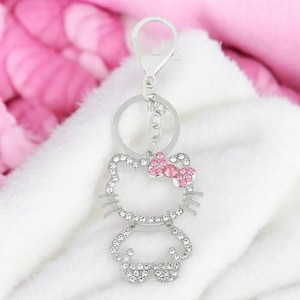 May include: A silver charm keychain featuring a rhinestone-encrusted Hello Kitty design with a pink bow. The keychain has a silver ring and a lobster claw clasp.