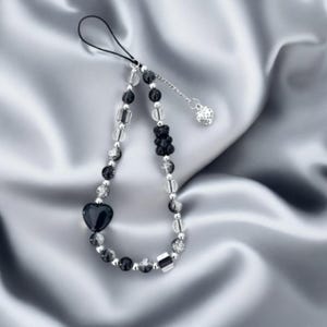 Puede incluir: Un charm de teléfono de cuentas negras y transparentes con un charm en forma de corazón y una cadena plateada. El charm está sobre un fondo de satén blanco.