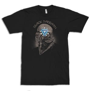 May include: Black t-shirt featuring the band name "Black Sabbath" above a detailed illustration. The illustration depicts a skeletal figure with a blue triangle in place of an eye. The design is in shades of gray and blue.