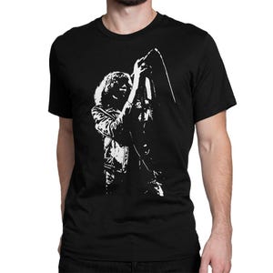 Puede incluir: Camiseta negra con un gráfico blanco de una persona sosteniendo un micrófono. El gráfico muestra a una persona con pelo largo usando una chaqueta. La camiseta está hecha de un material suave.