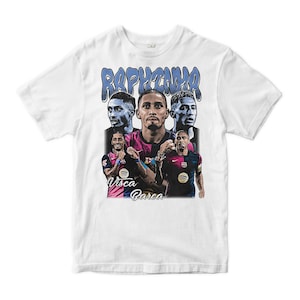 Può includere: Una t-shirt bianca a maniche corte con una stampa grafica del calciatore Raphinha in diverse pose e ritratti. Il design include il testo RAPHINHA in lettere a bolla blu nella parte superiore e Visca Barca in corsivo nella parte inferiore.