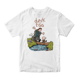 以下が含まれることがあります： 白いTシャツ。漫画のイラストで、丸太の上で小川を渡る2人のキャラクターが描かれています。グラフィックには、茶色の文字で「dunk and Egg」というテキストが含まれています。キャラクターは気まぐれなスタイルで描かれています。